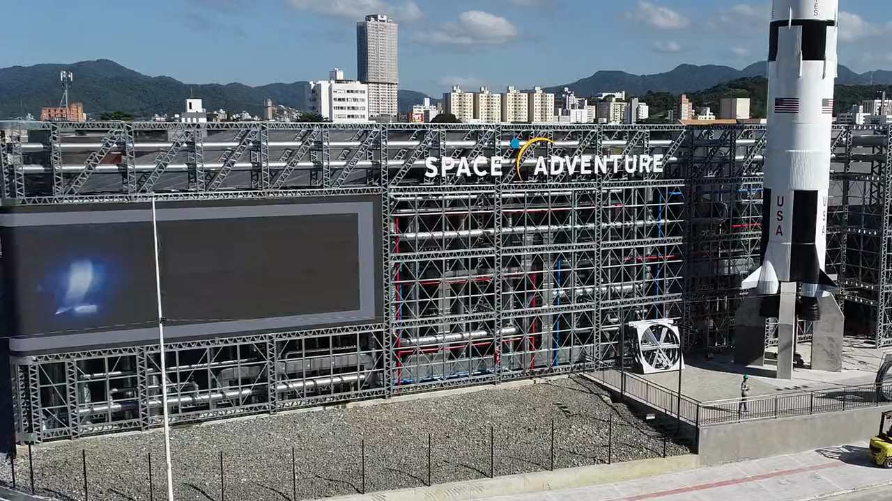 Fachada do Space Adventure, o Museu da NASA em Balneário Camboriú: malha
            estrutural em steel frame contra o céu, com a réplica do foguetão Saturn V ao
            lado.